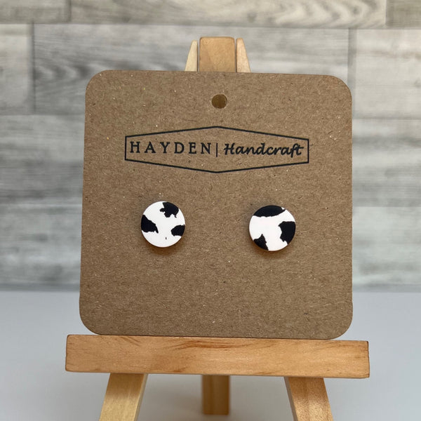 Pair of circular black and white cow print earrings on a Hayden Handcraft branded card with a wooden stand against a wooden panel background.
