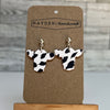 Cow print earrings on a Hayden Handicraft card with a wooden stand against a wooden panel background.