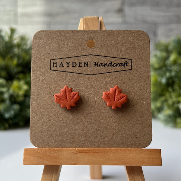 Red maple leaf earrings on a Hayden Handcraft card with a wooden stand against a gray brick wall.